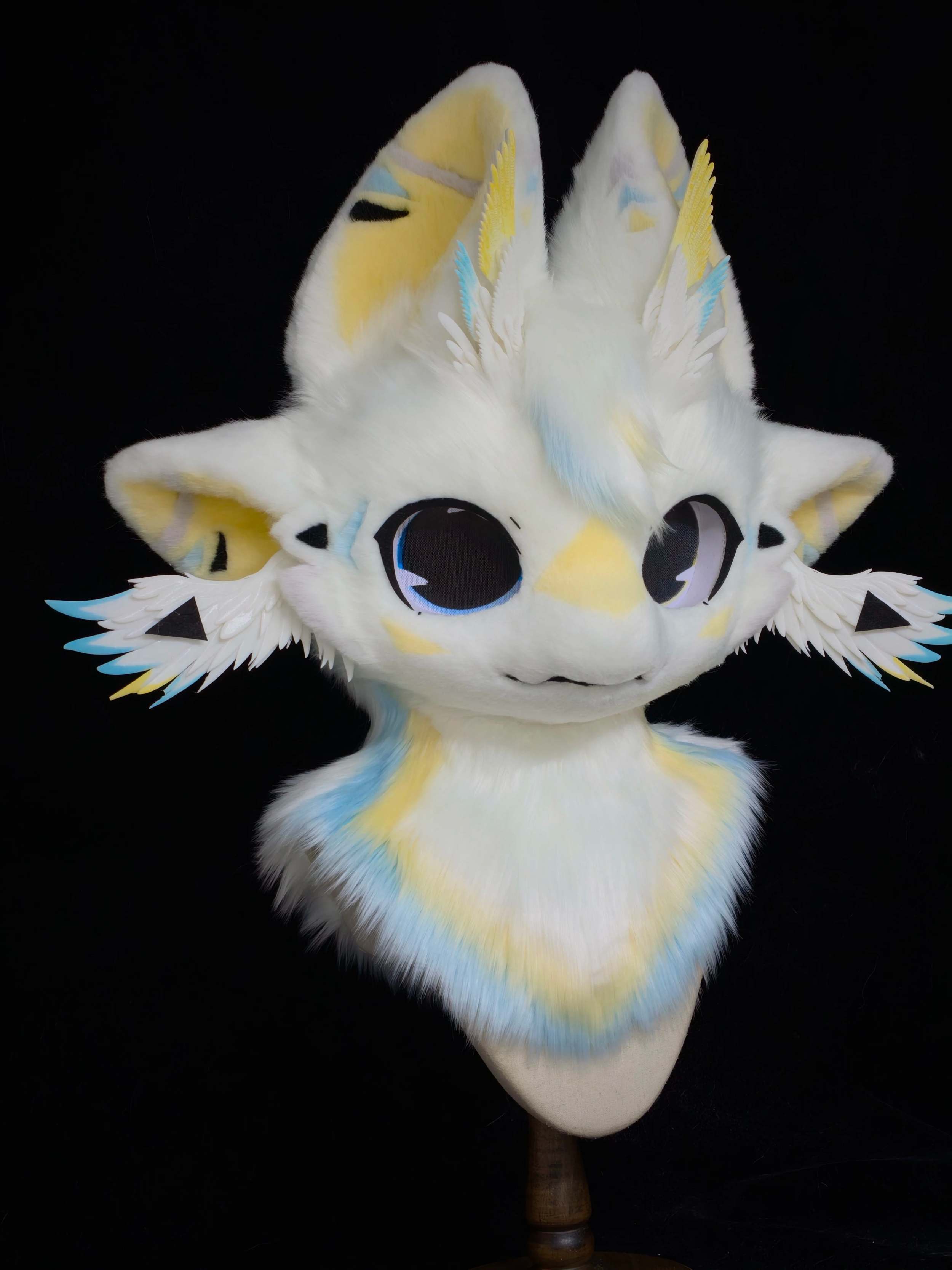 Handmade white and yellow fursuit head with blue accent feathers and large dark eyes, displayed on a stand