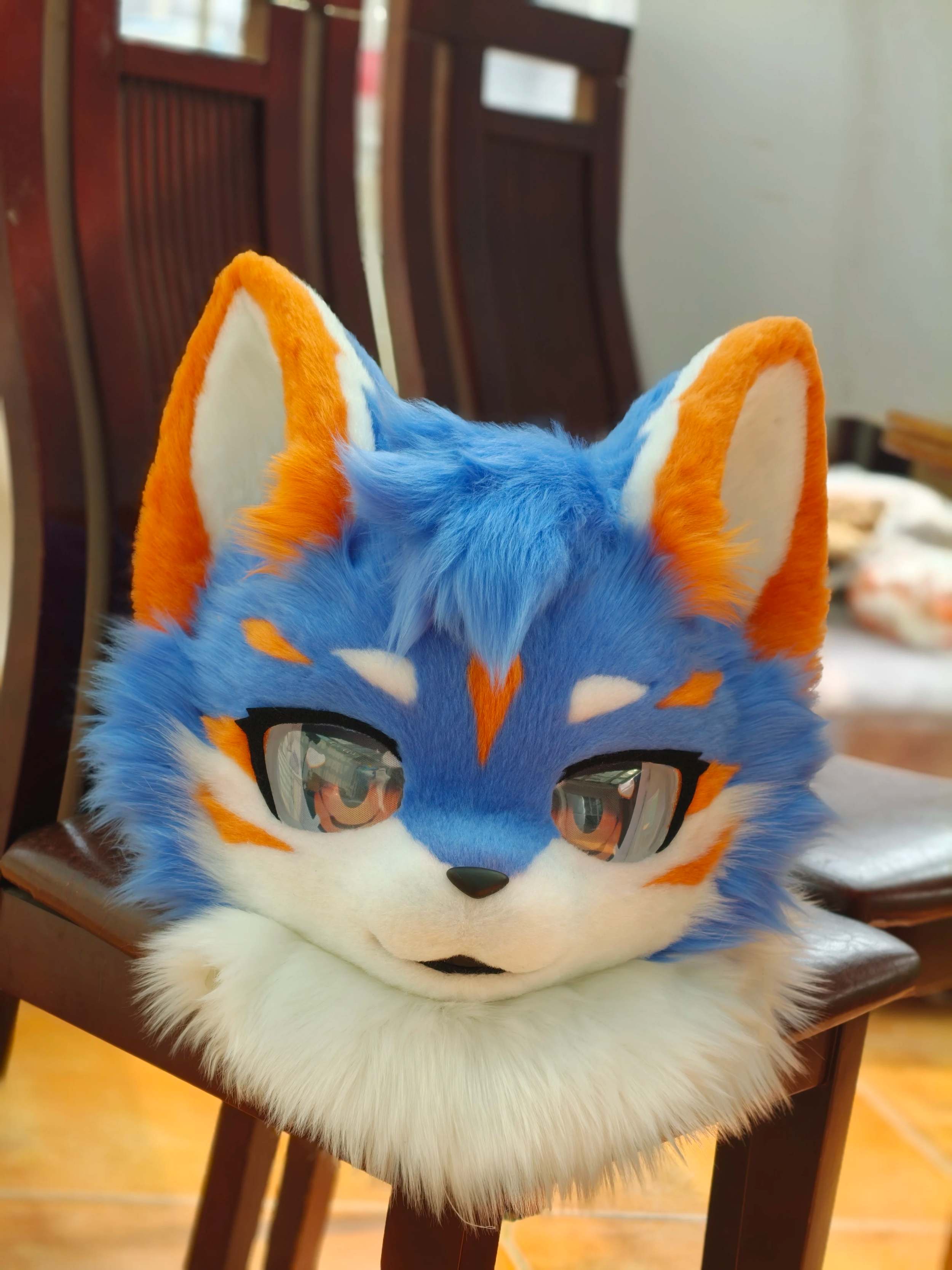 Blue and orange kemono fox fursuit head with reflective slit eyes and white chin ruff, resting on chair